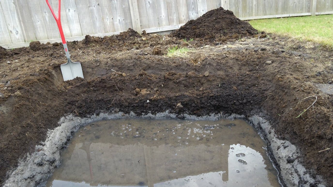 Gegraven put in een Nederlandse achtertuin waarin de bodemlagen zichtbaar zijn: donkere teelaarde, een zwarte veenband en grijze klei, met grondwater dat onderin de put staat