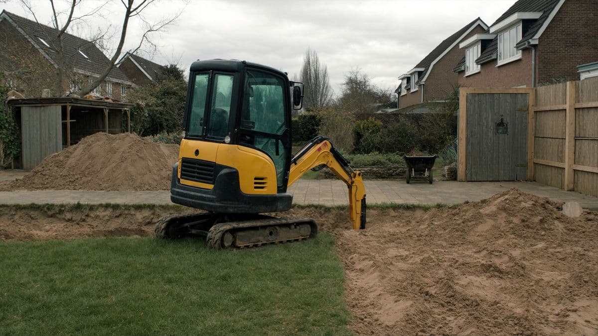 Een minigraafmachine graaft een put in een Nederlandse achtertuin met schutting en zandhopen