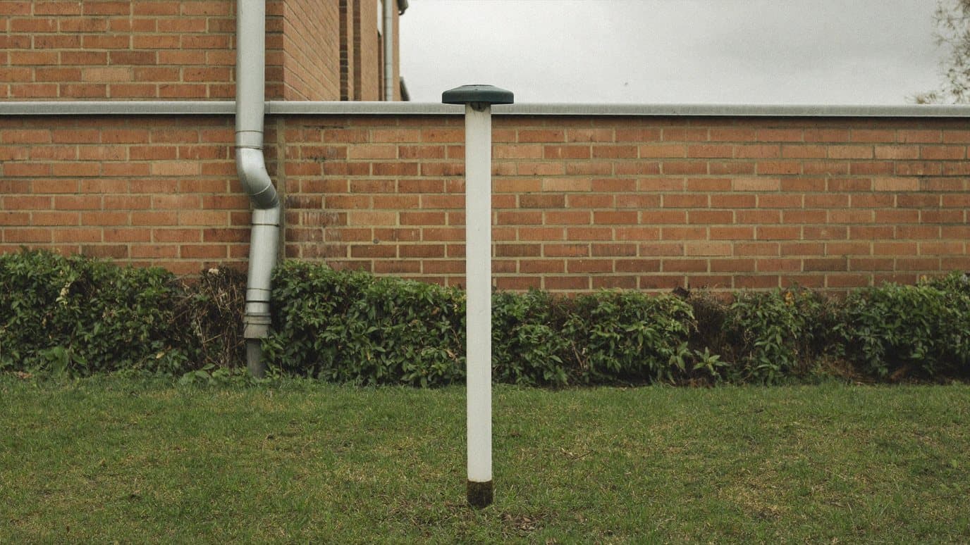 Witte peilbuis met donkere dop in een grasstrook langs een Nederlandse bakstenen woning, waarmee de grondwaterstand wordt gemeten